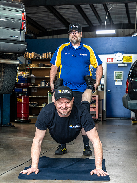 Two men in a garage, one doing a push-up with the other standing on his back, smiling.