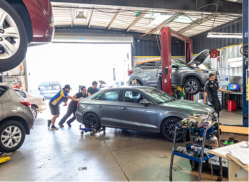 People pushing a gray car on a dolly in an auto repair shop. Other cars are on lifts.