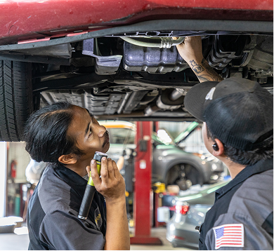 Two mechanics inspecting the underside of a car in a garage, one using a flashlight.