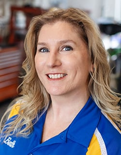 Woman with blonde hair, smiling at the camera, wearing a blue and yellow uniform in a workshop setting.