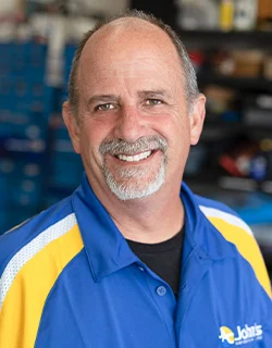 Man with graying goatee smiling, wearing a blue and yellow collared shirt in a shop.