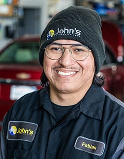 A smiling mechanic wearing a black beanie and John's Automotive uniform, in front of a car.