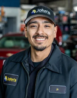 Smiling man in John's auto service uniform, wearing a cap. He is in front of a car in a garage.