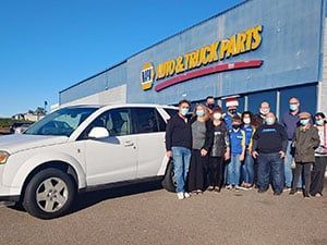 Group of people stand in front of a NAPA Auto & Truck Parts store with a white SUV.