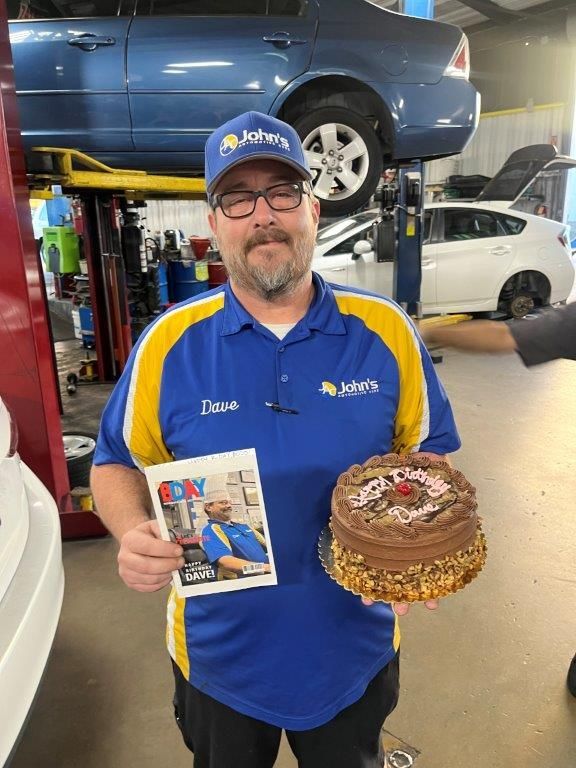 Man in John's Auto Service uniform holding a magazine with his photo and a birthday cake in an auto repair shop.