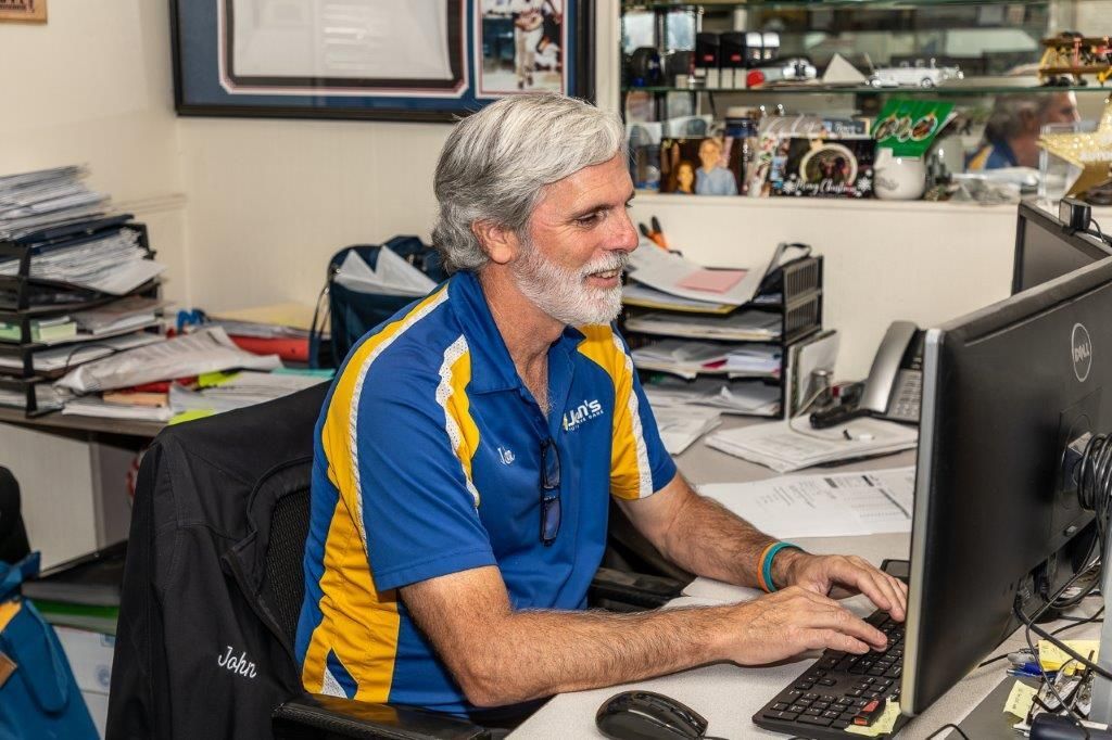 Man with gray hair and beard typing at a desk with a computer. He wears a blue and gold polo shirt.