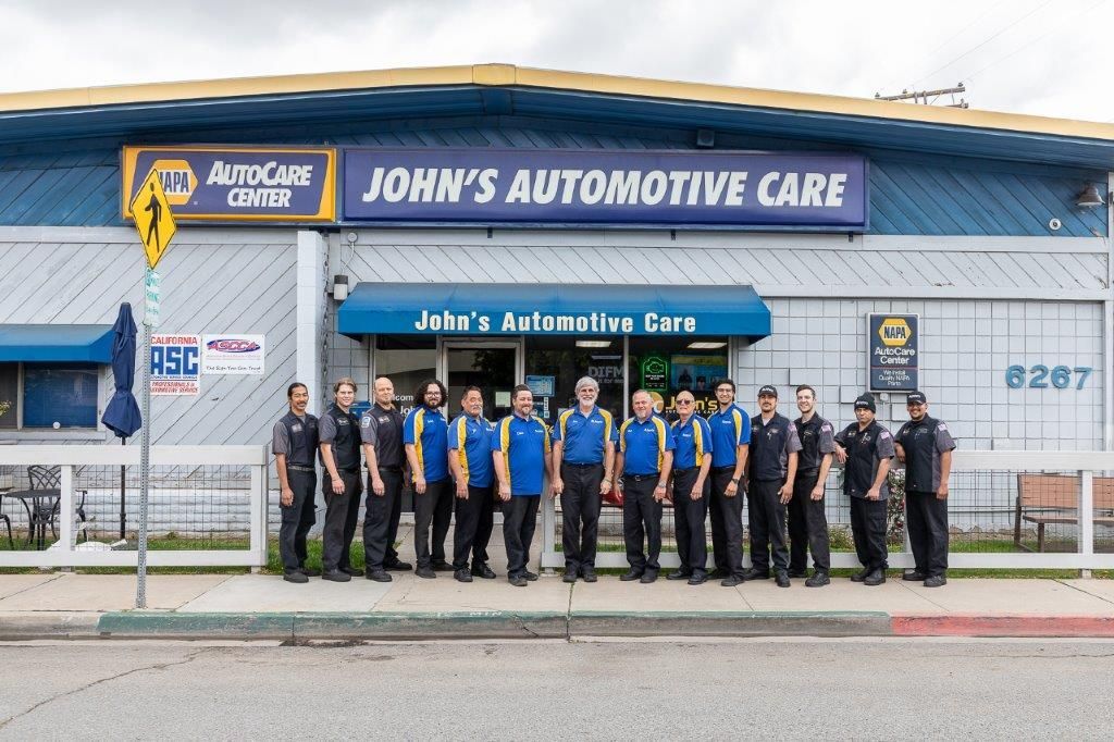 Group of people in blue and black shirts stand in front of John's Automotive Care building.