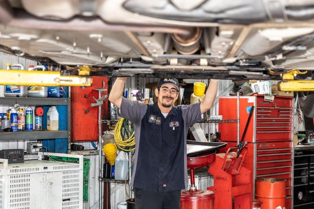 Mechanic working under a car, smiling, in a shop. Wearing work clothes, tools visible.