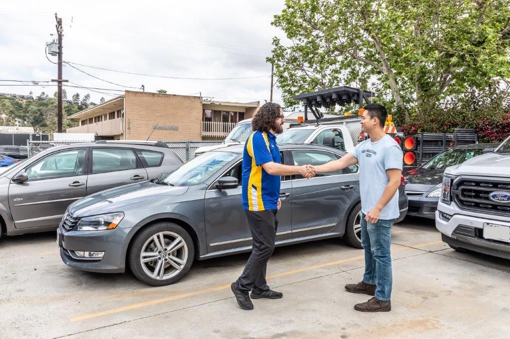 Two men shaking hands in a parking lot next to a gray car. One wears a blue and yellow shirt, the other a light blue t-shirt.