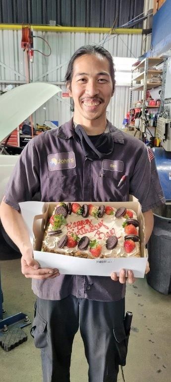 Man in mechanic uniform smiling, holding a birthday cake in a garage.