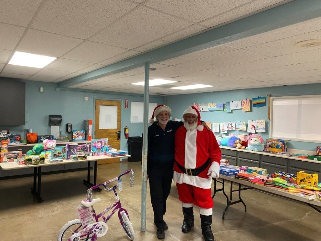 Two people in Santa hats pose by tables of toys in a room.