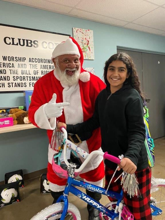 Santa points at a girl holding a new blue bicycle. They smile in a room with a club sign.