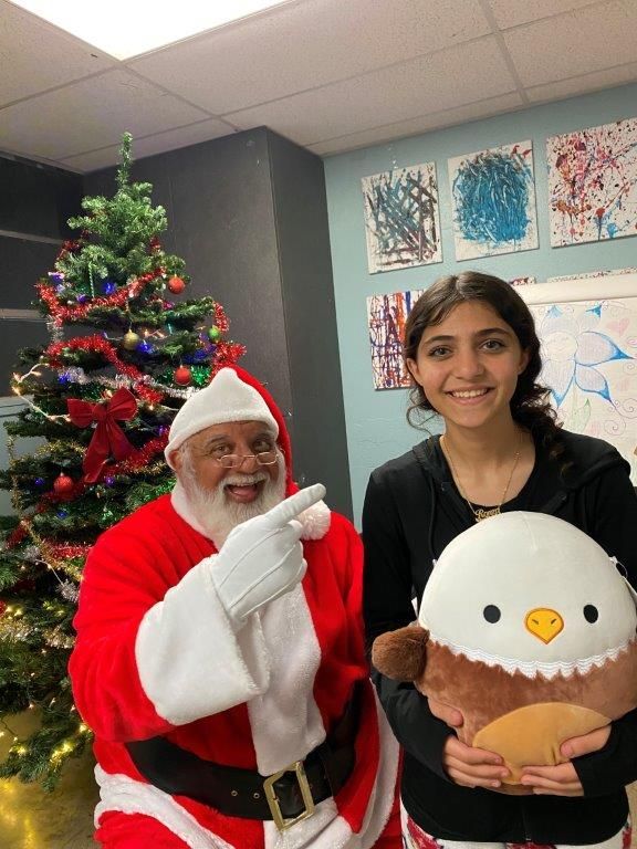 Santa points at a smiling person holding an eagle plush, next to a decorated Christmas tree.