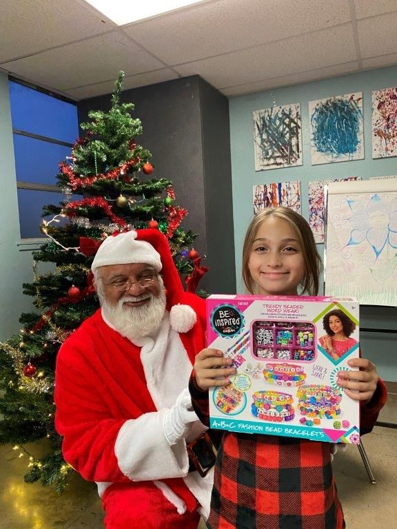 Santa with a girl holding a craft kit in front of a Christmas tree.