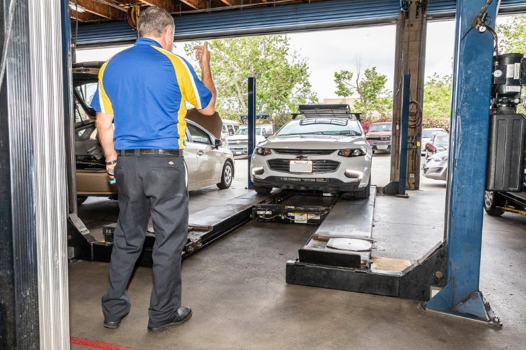 Mechanic directing a white car onto a lift in a garage. He wears a blue and yellow shirt.