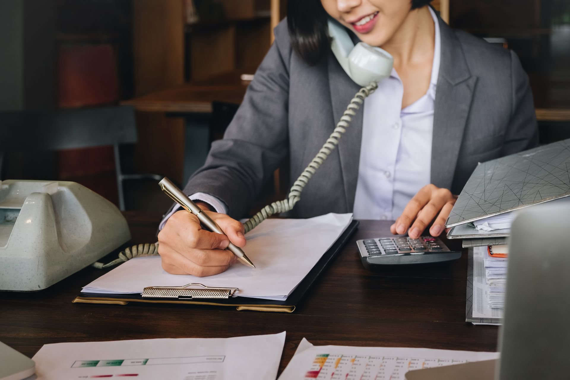 Woman in gray suit, using landline phone, writing on clipboard and calculating. Desk setting.