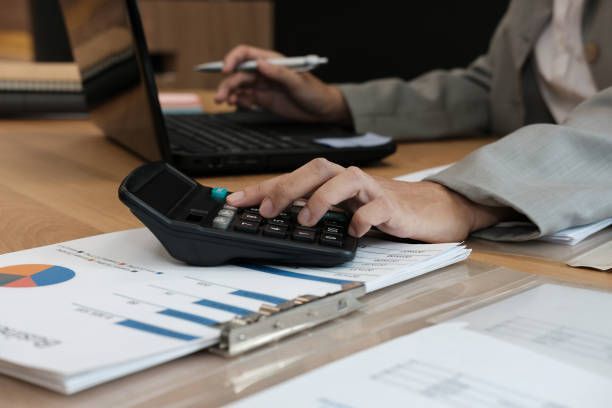Person calculating on a calculator while working at a laptop and reviewing financial documents.