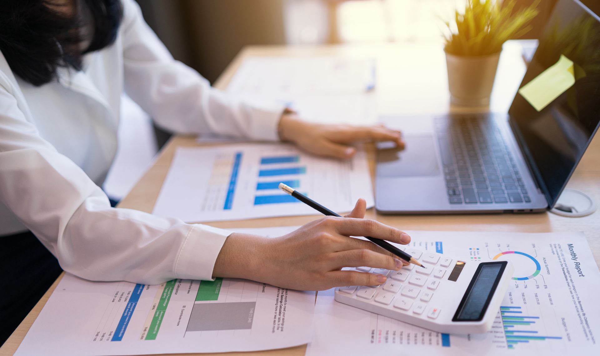 Person using calculator and laptop, reviewing charts and graphs at a desk, sunlight in background.