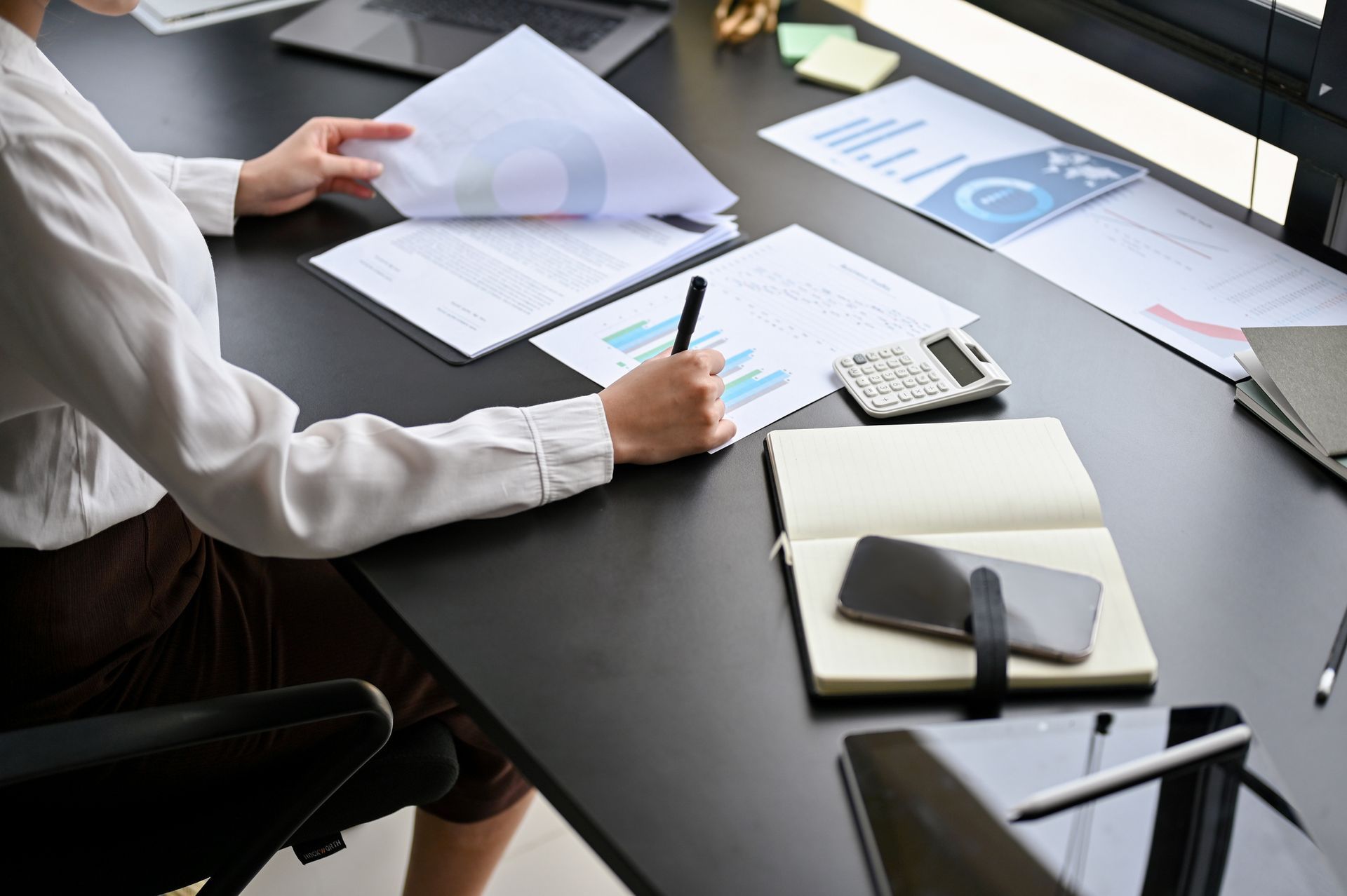 Person at desk reviewing papers, taking notes, using a calculator, and working on a laptop.