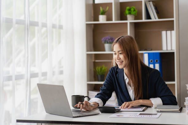 Woman in navy blazer working on laptop at a desk, smiling. White office with bookshelves and window.