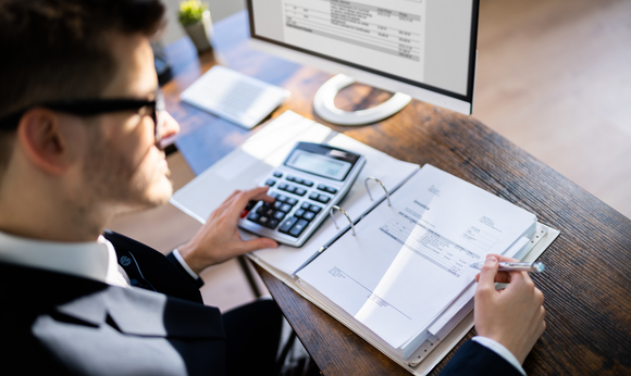 Man in glasses uses calculator and reviews documents at a desk with a computer displaying a document. Man in glasses uses calculator and reviews documents at a desk with a computer displaying a document.