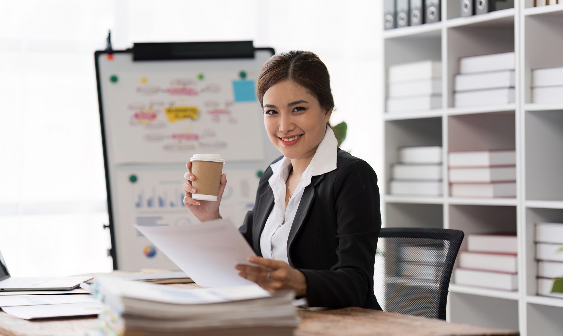Woman in a blazer holds coffee, reviewing papers at desk in office; whiteboard and shelves in background.