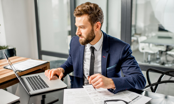 Man in suit working on laptop at desk, documents and glasses nearby. Man in suit working on laptop at desk, documents and glasses nearby.