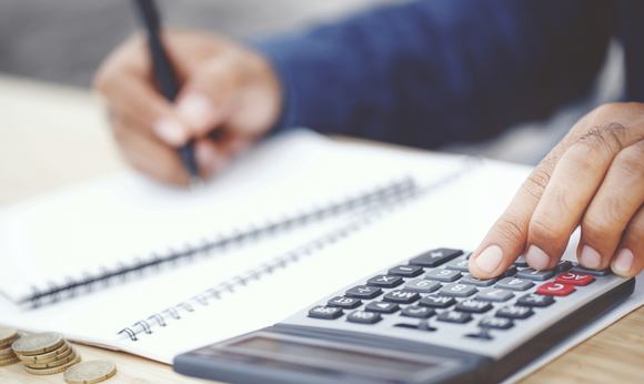 Person using a calculator and writing in a notebook, next to stacked coins on a wooden surface. Person using a calculator and writing in a notebook, next to stacked coins on a wooden surface.