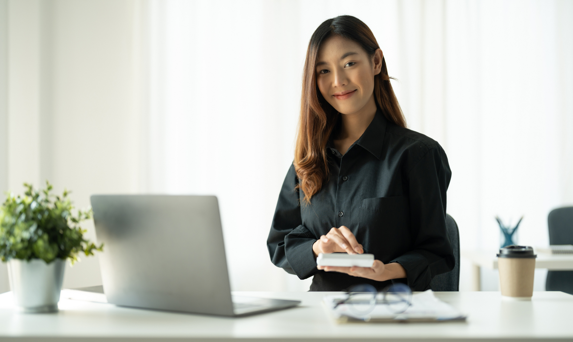Woman in black shirt uses a calculator at a desk with laptop, plant, and coffee. Woman in black shirt uses a calculator at a desk with laptop, plant, and coffee.