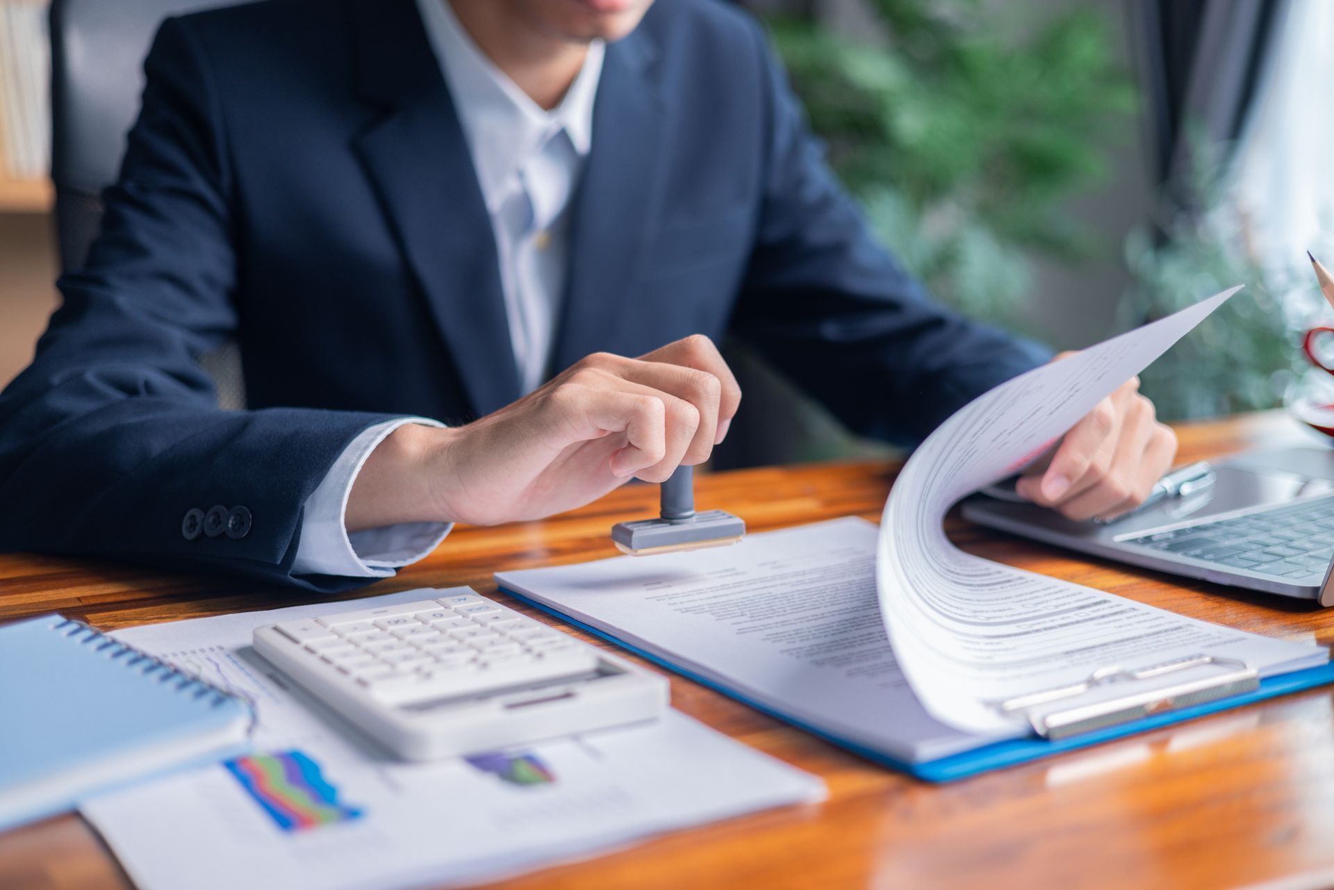 Person in suit stamping document with papers, calculator, and laptop on a desk.