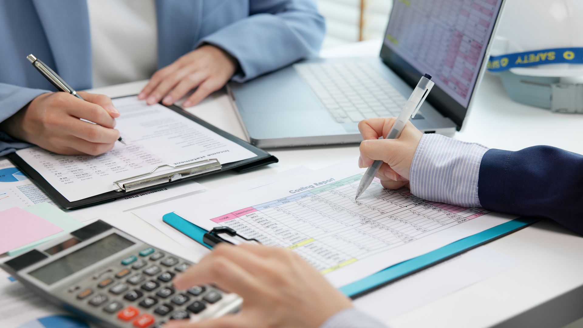 People working at a desk, writing on documents, using a calculator, and working on a laptop.