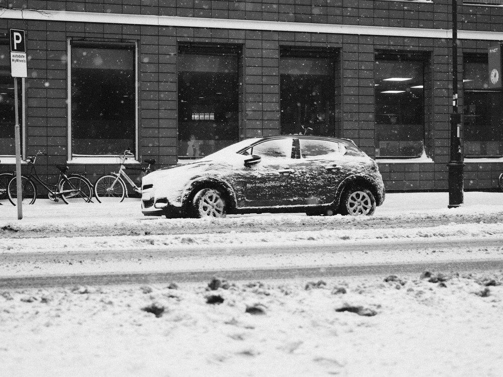 A side profile, black-and-white photograph of a snow-dusted compact car parked along a slushy city street. In the background, a large tiled building features tall glass windows, with a couple of bicycles parked to the left next to a public sign post.