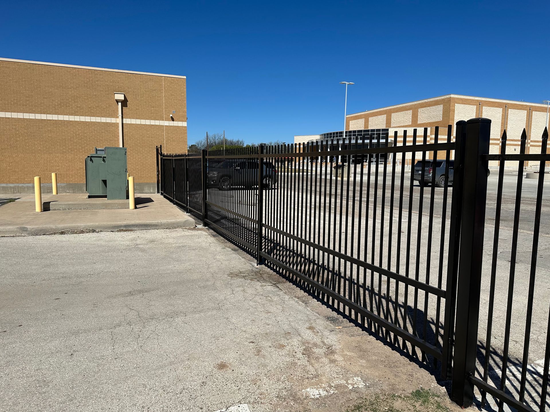 A black metal fence surrounds a parking lot with a brick building in the background.