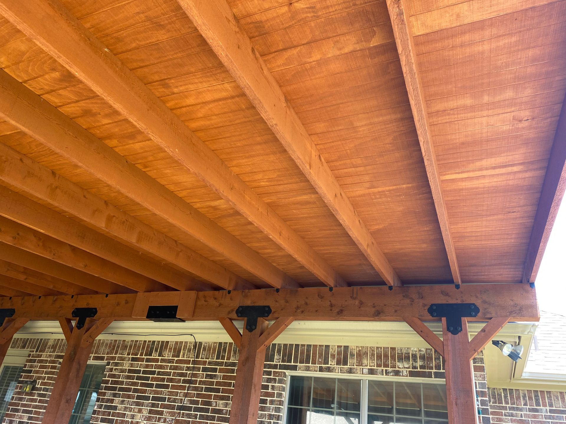 The ceiling of a wooden pergola with a brick wall in the background.
