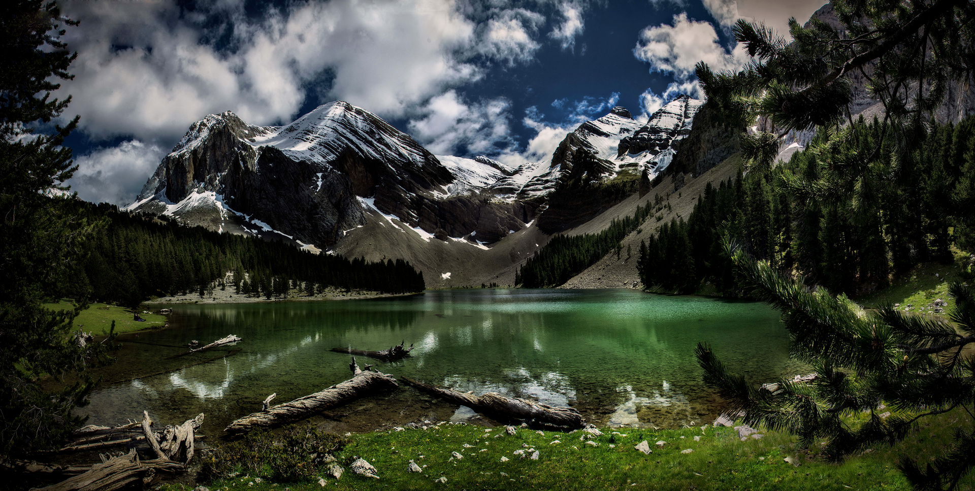 Escena de un lago de montaña con picos nevados, exuberantes árboles verdes y cielo azul nublado.