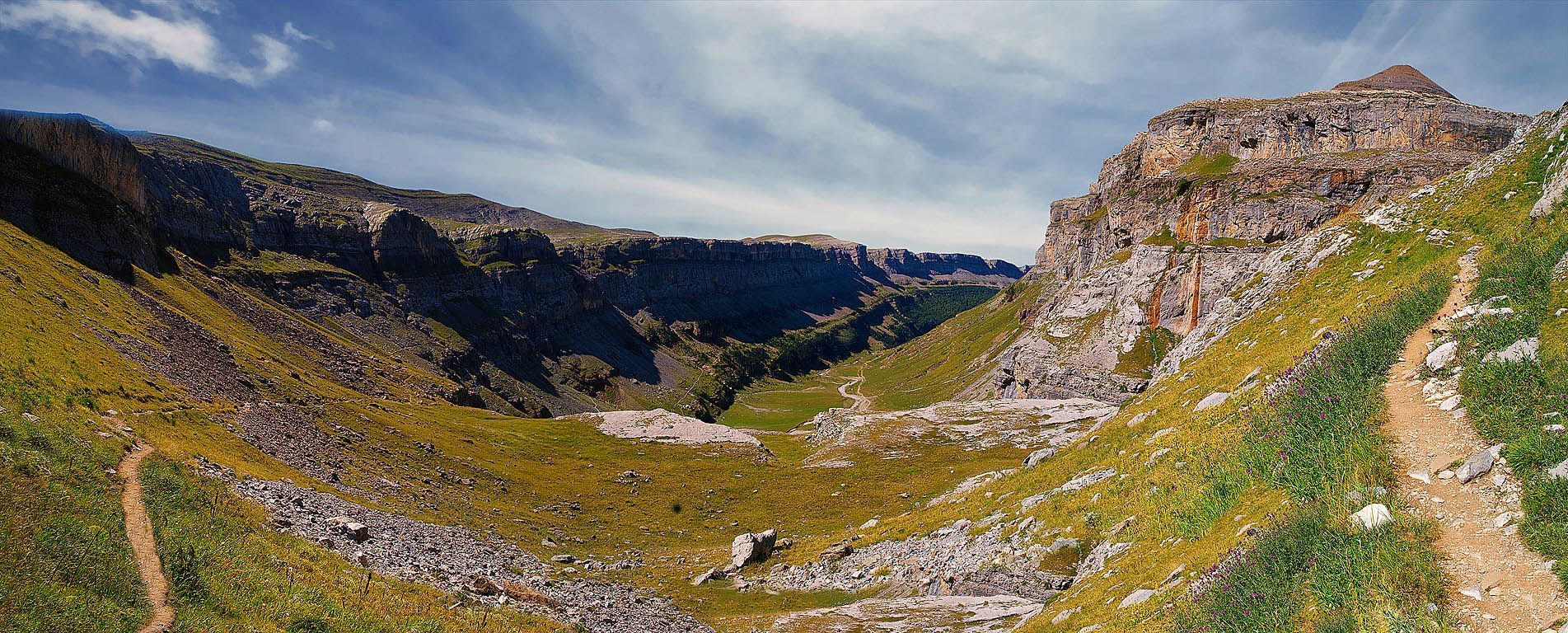 Vista panorámica de un valle de montaña con un sendero sinuoso. Hierba verde, terreno rocoso y acantilados bajo un cielo azul