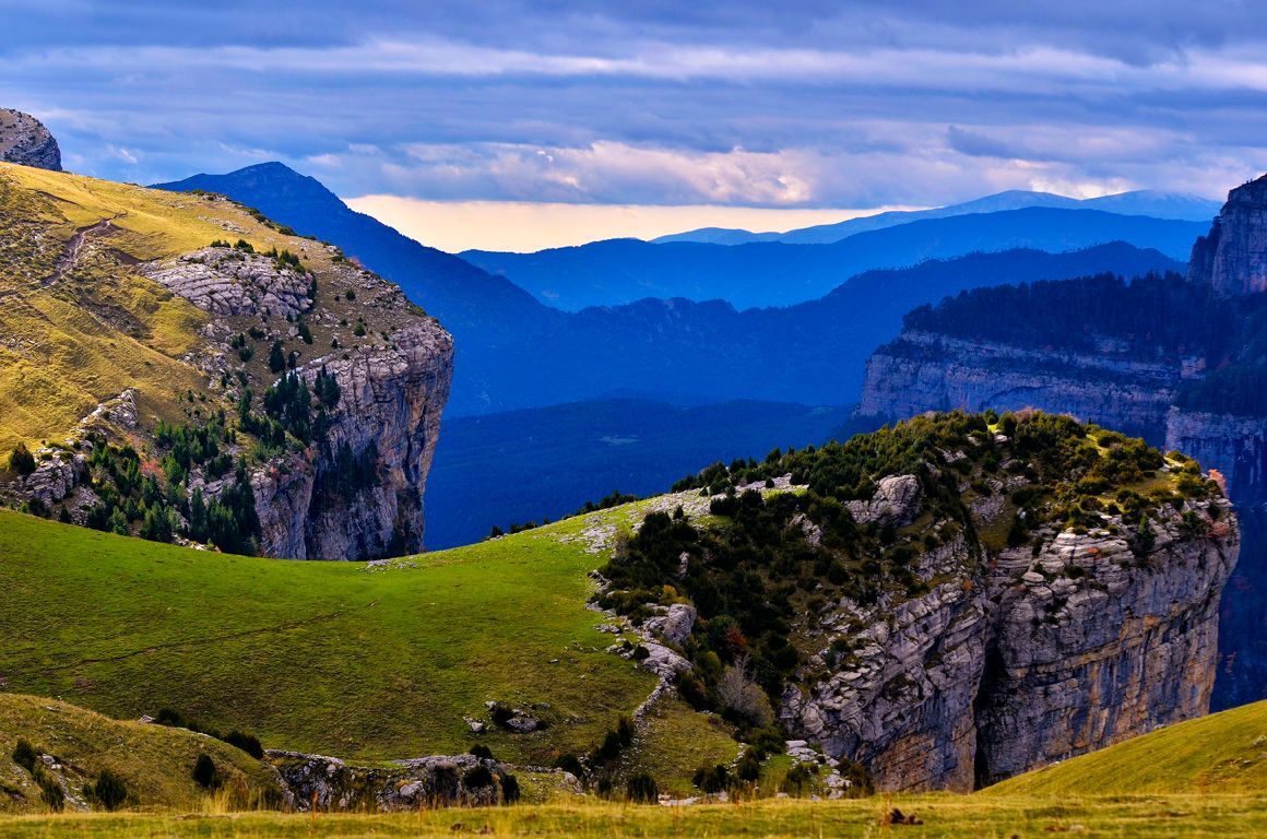 Laderas verdes y cubiertas de hierba, enmarcadas por acantilados rocosos, ofrecen una vista de cadenas montañosas azules.