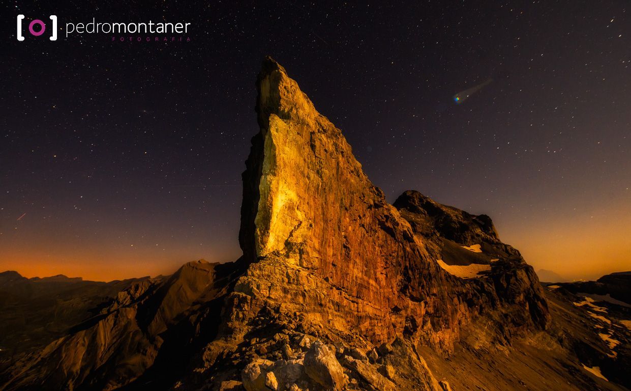 Montaña irregular iluminada por la luz, cometa cruzando un cielo nocturno estrellado.