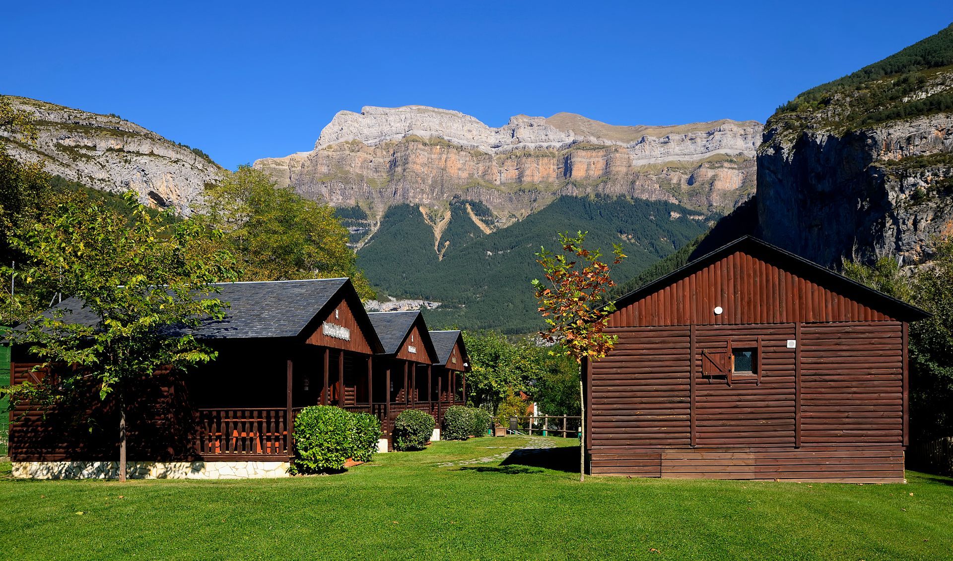 Cabañas de madera sobre un césped con montañas al fondo bajo un cielo azul despejado.