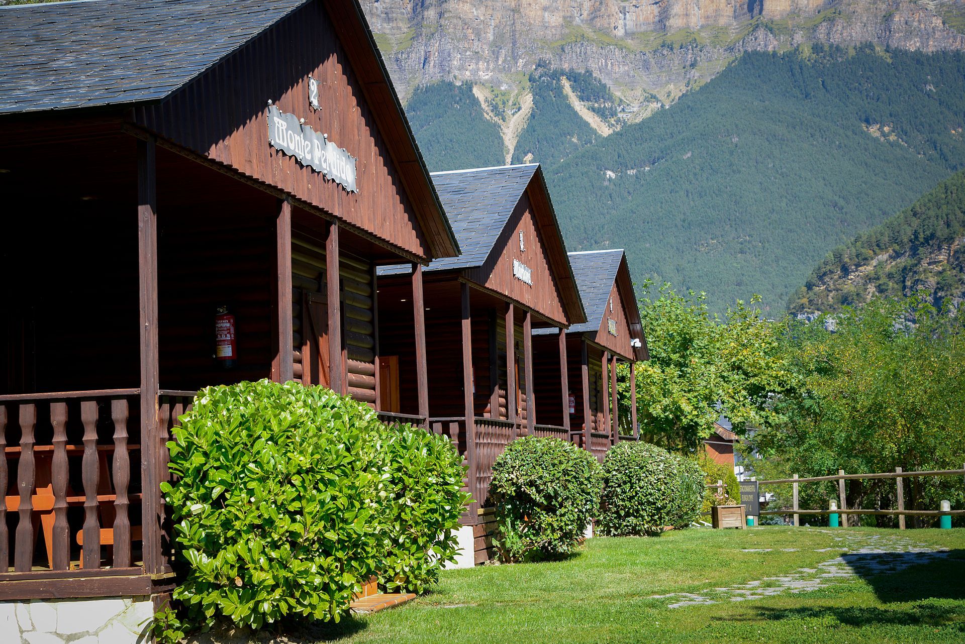 Cabañas de madera con techos oscuros y montañas de fondo. Arbustos y césped de un verde exuberante.