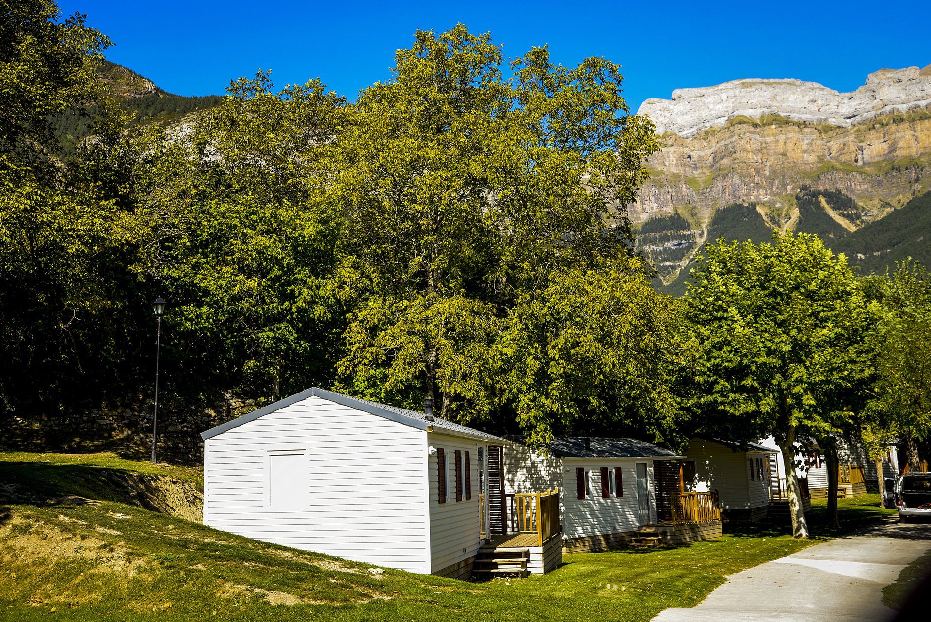 Cabañas en una zona verde con césped, árboles y montañas al fondo, bajo un cielo azul.