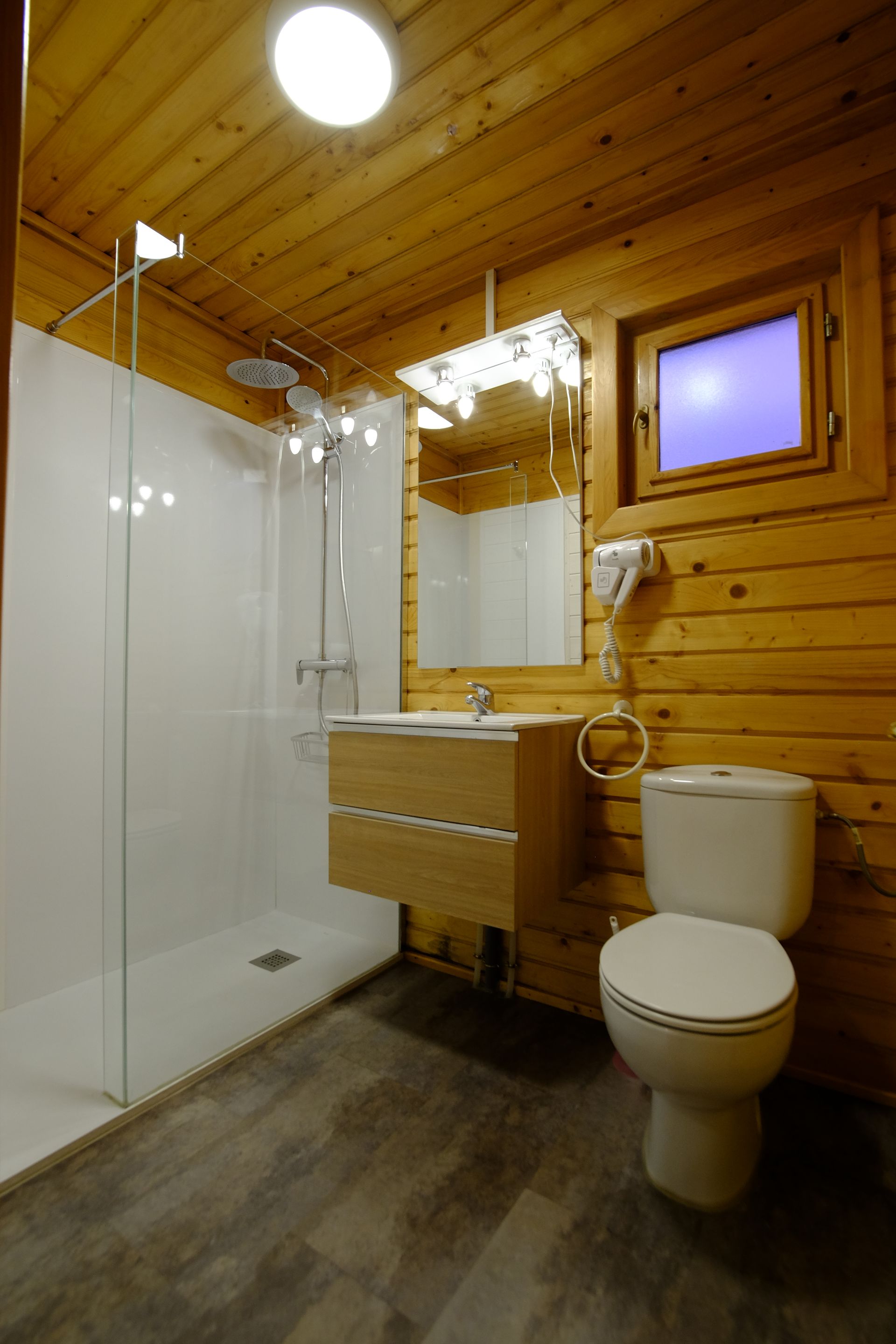 Baño con paneles de madera, ducha, inodoro y lavabo. Ventana con luz azul tenue.