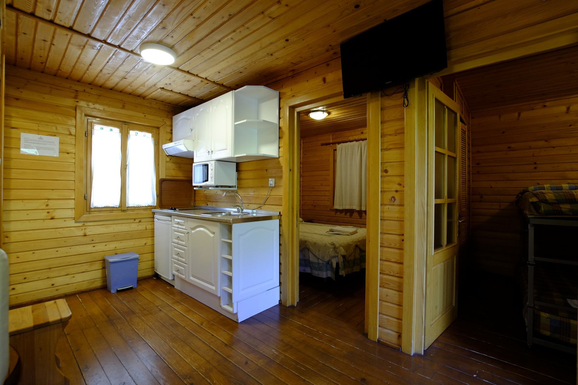 Interior de una cabaña de madera con cocina, puerta a un dormitorio y televisor.