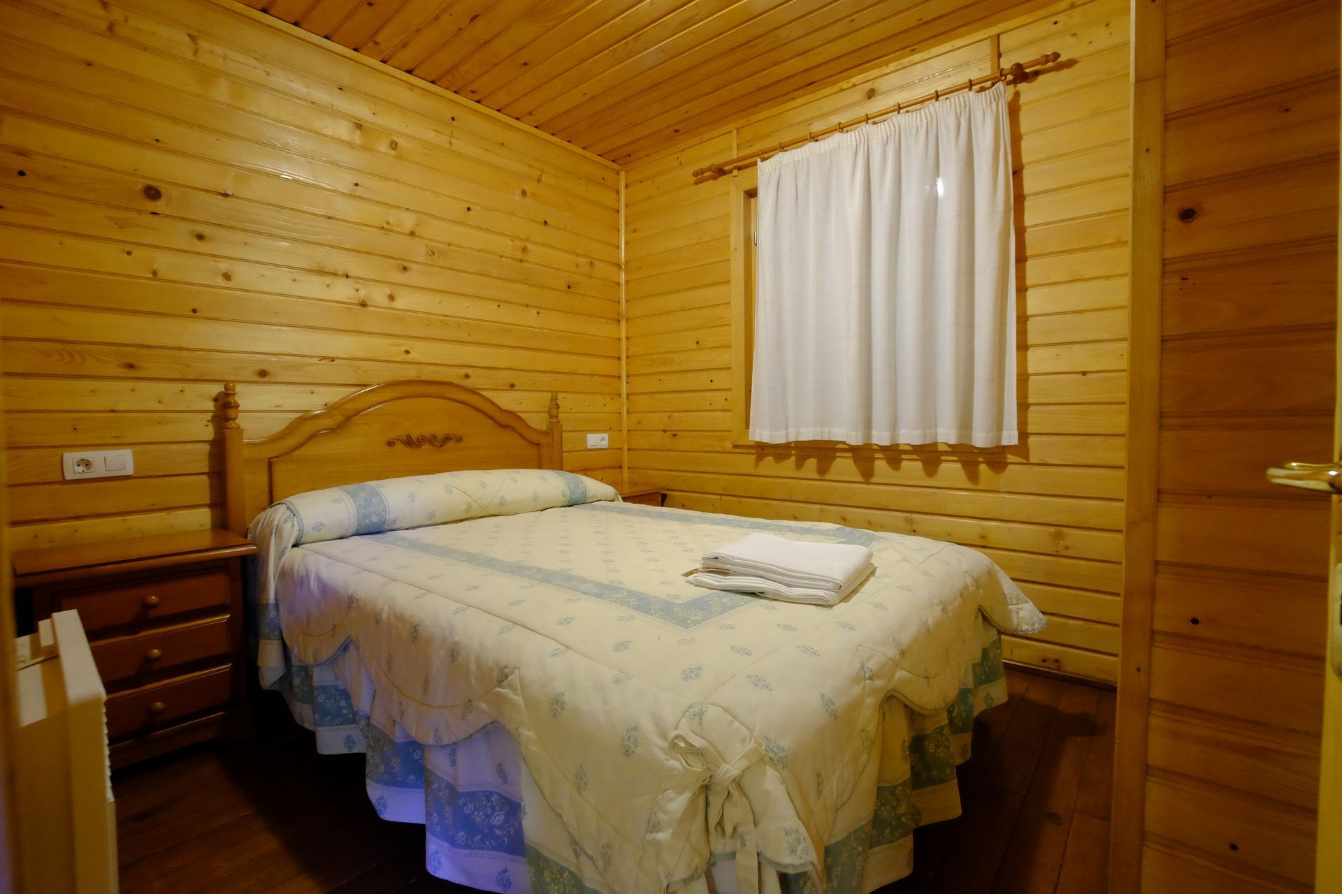 Dormitorio con paneles de madera, una cama doble con edredón blanco y una ventana con cortina blanca.
