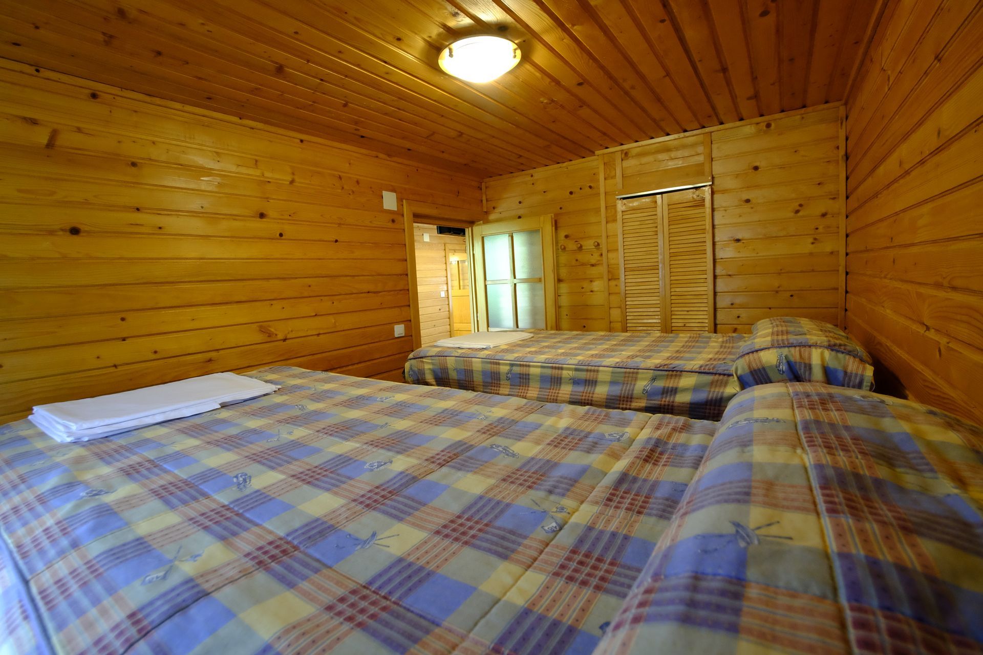 Dos camas en una cabaña con paneles de madera. Colchas a cuadros, puerta trasera cerrada.