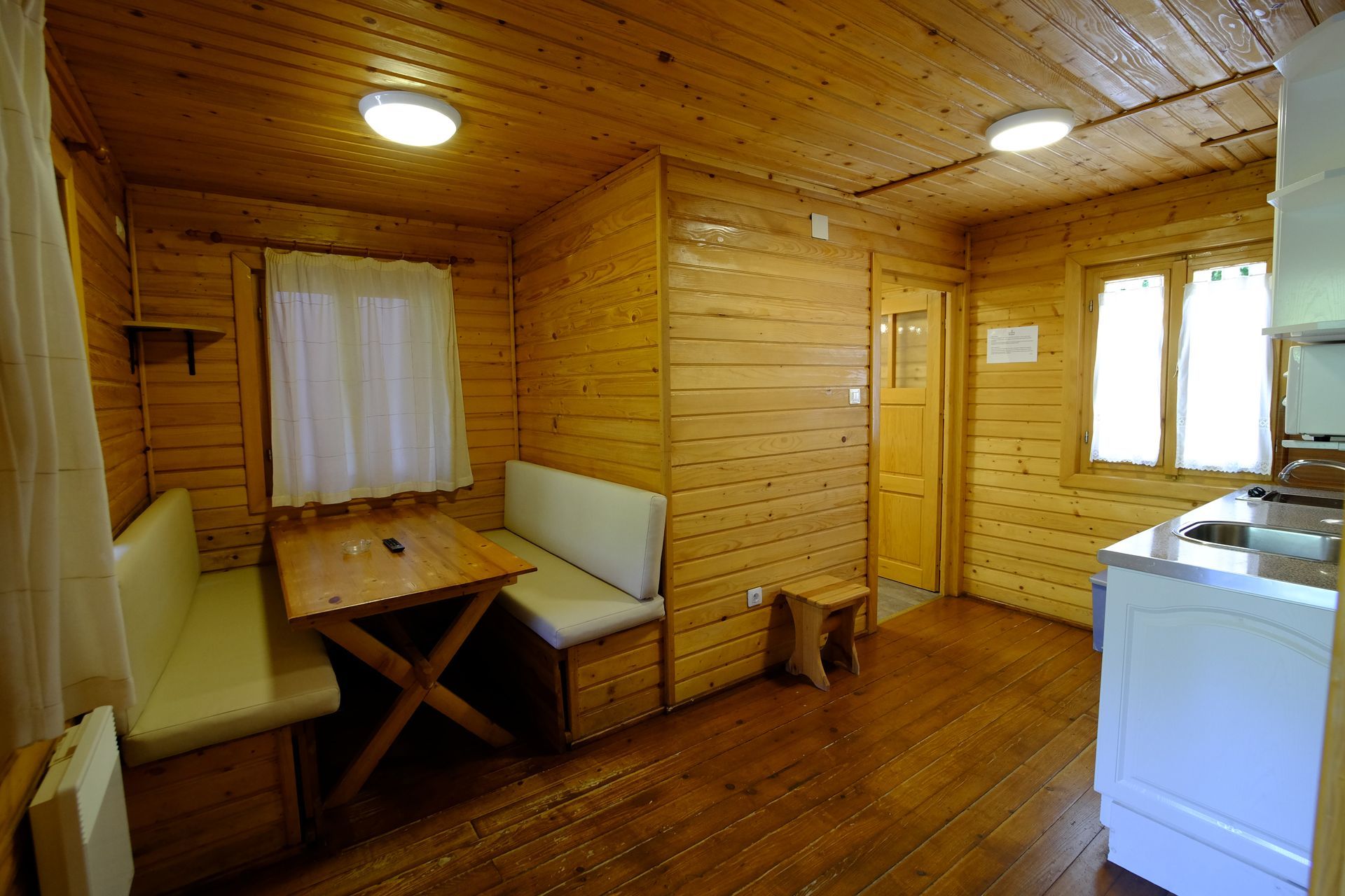 Interior de una pequeña cabaña de madera con mesa, bancos y zona de cocina.