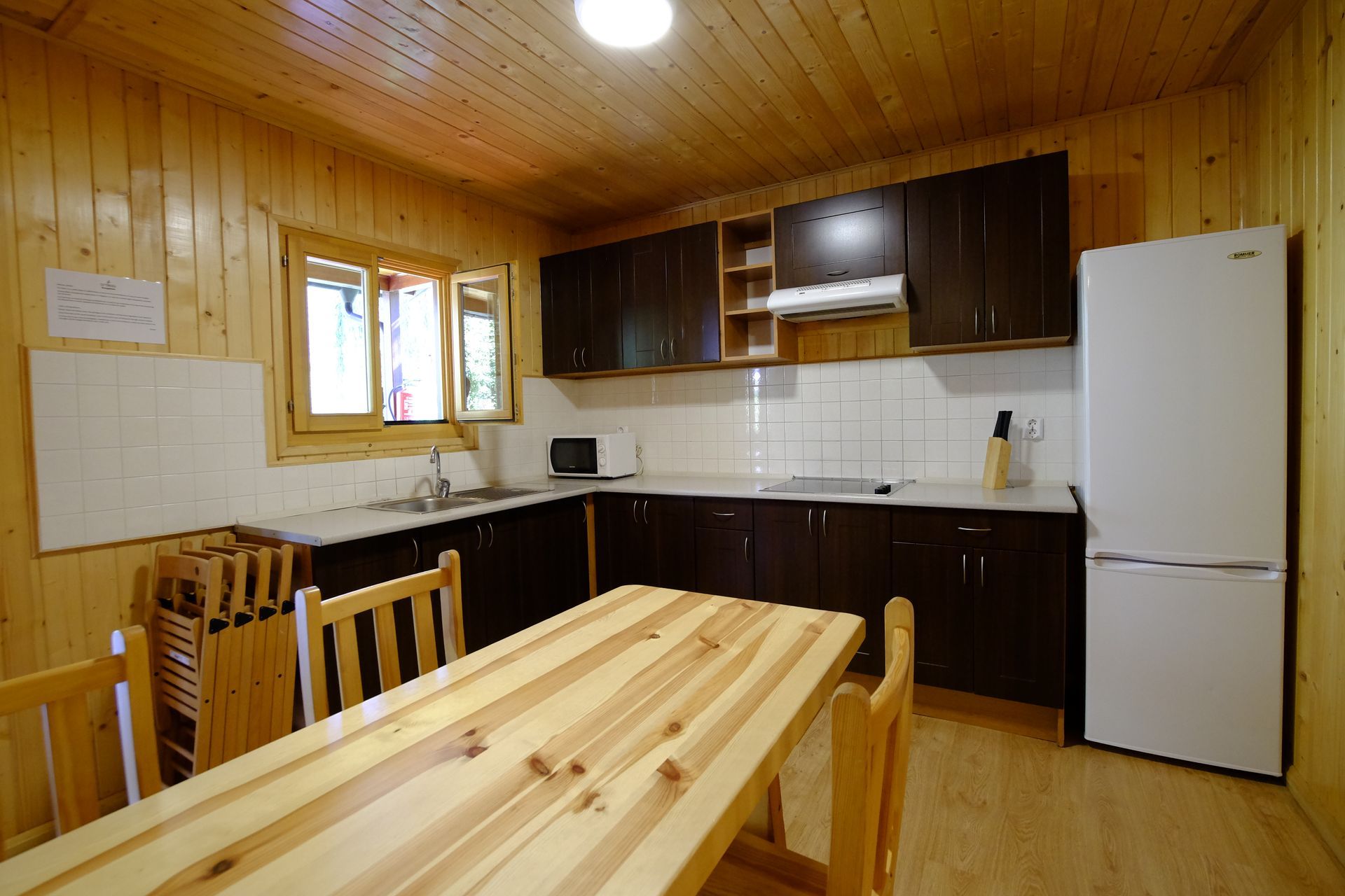 Cocina con paneles de madera, armarios oscuros, frigorífico blanco y mesa y sillas de madera clara.