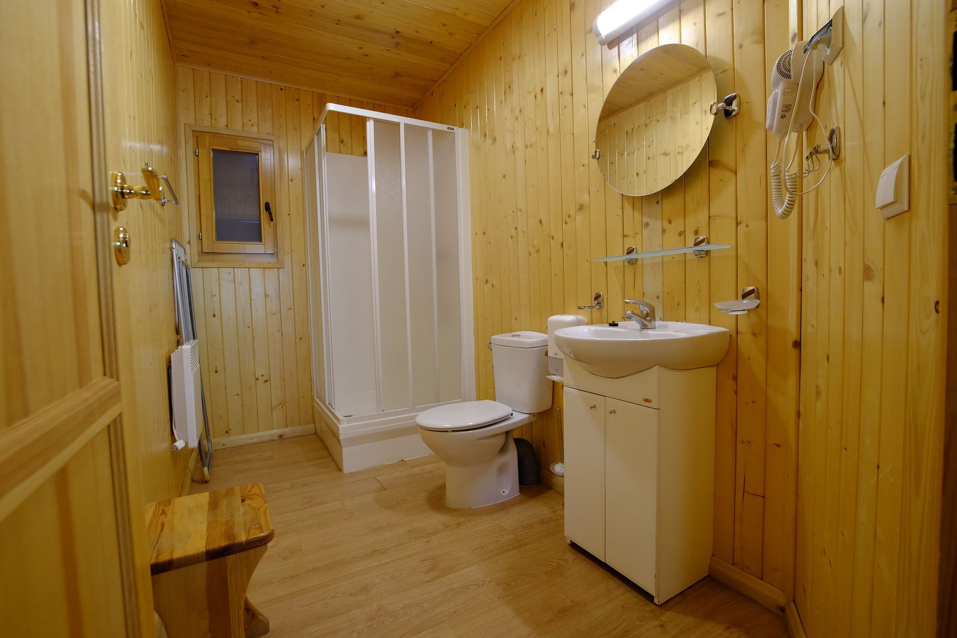 Baño con paneles de madera clara, cortina de ducha blanca, inodoro, lavabo y espejo redondo.