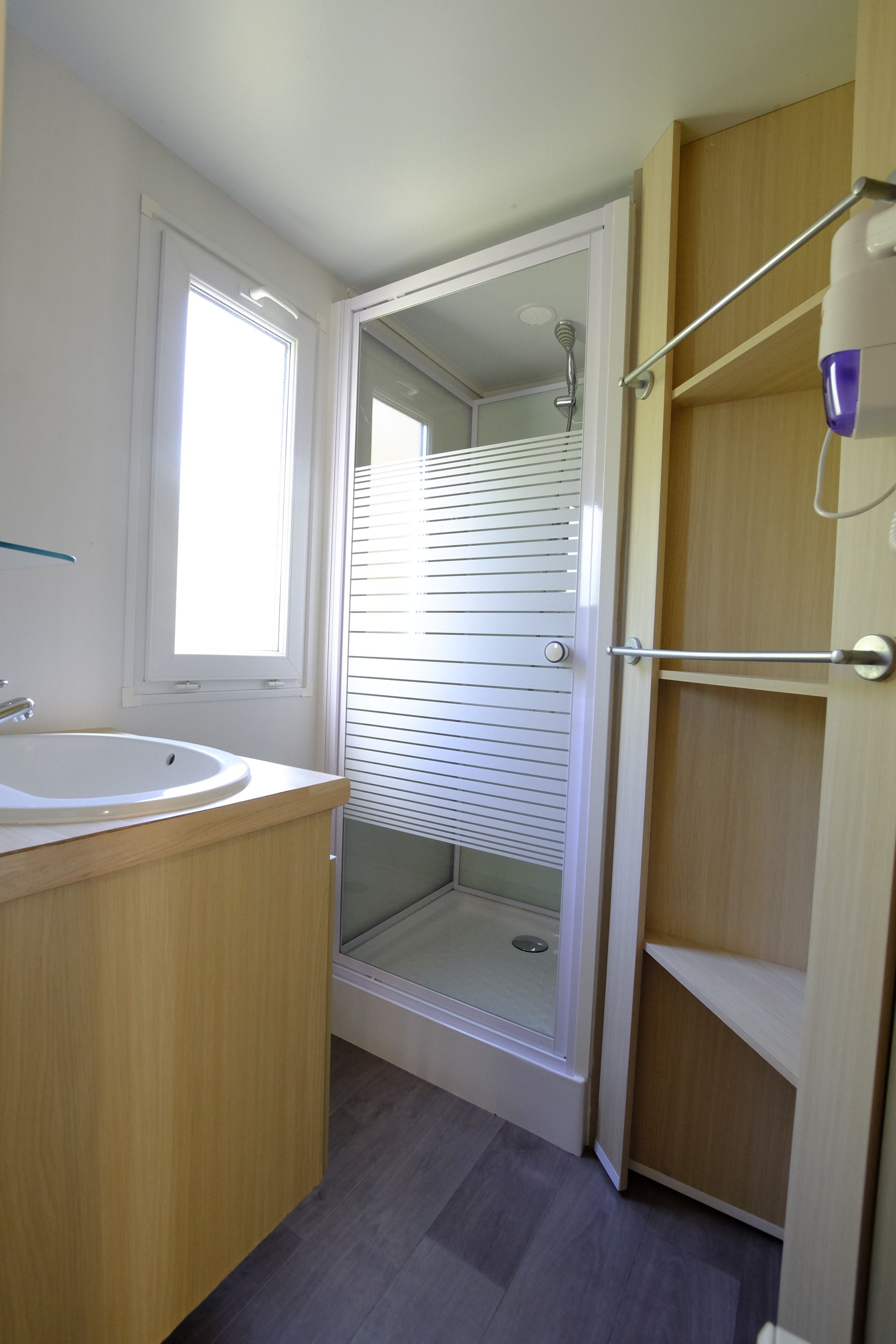 Baño con ducha, lavabo y estantes. Armarios de madera clara, suelo gris, mampara de ducha esmerilada.