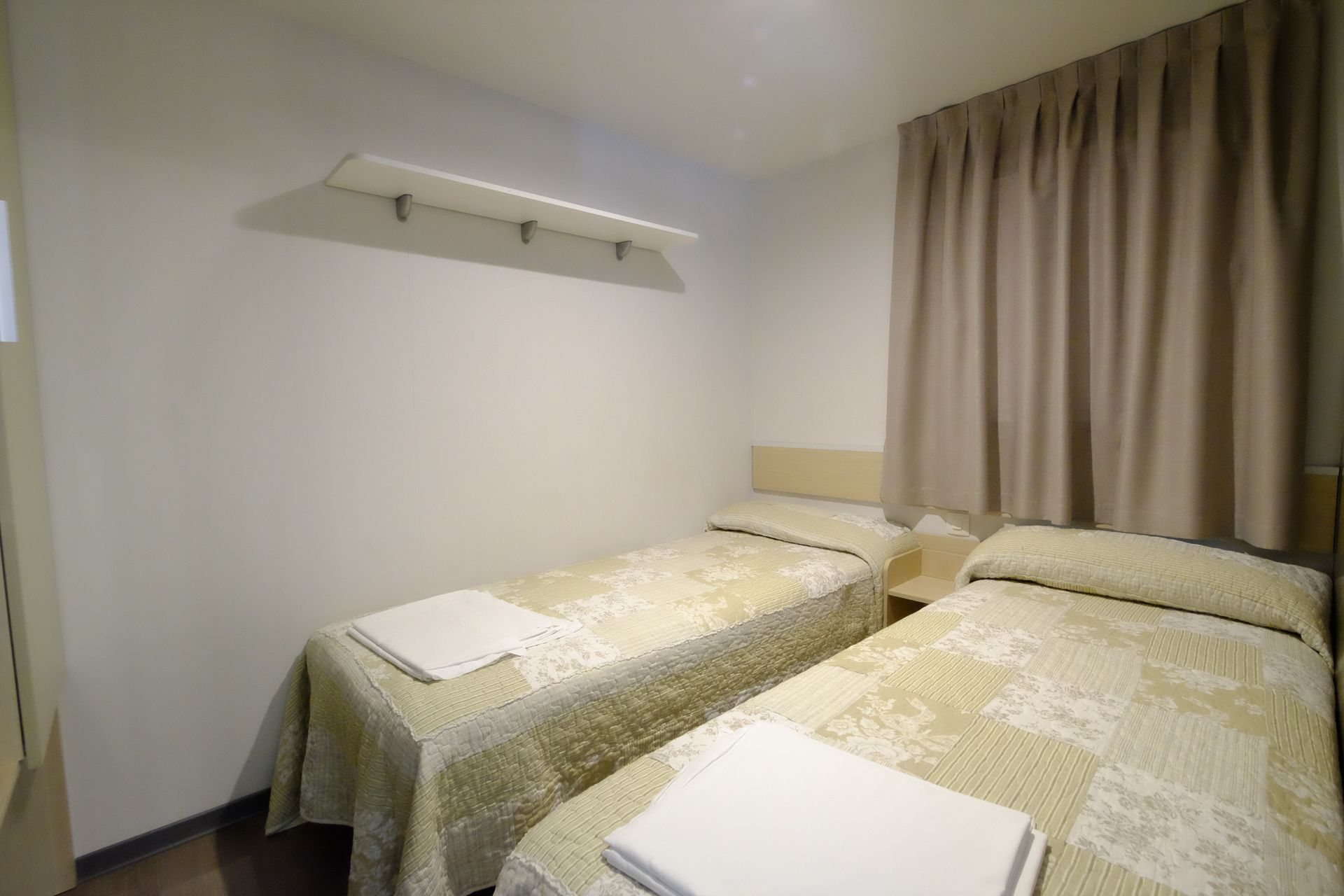 Dos camas individuales en una habitación pequeña con ropa de cama color canela, cortina beige y un estante.